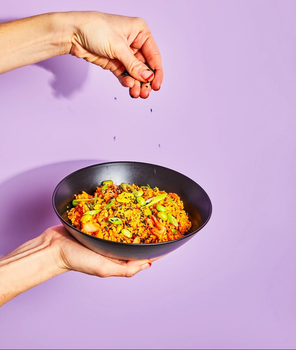 Hand holding a black bowl of grain salad against a lavender backdrop